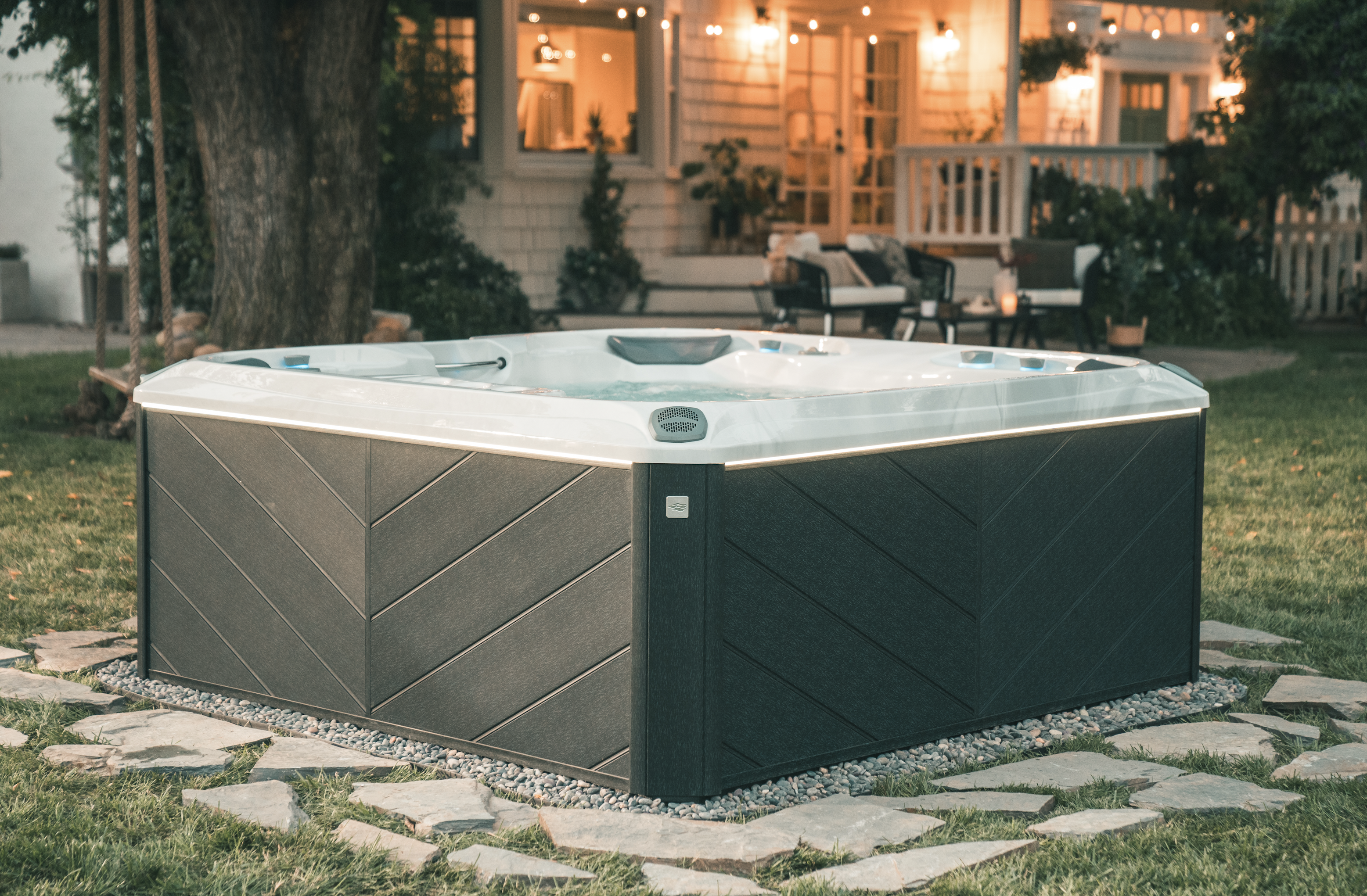 Hot tub in a backyard landscape in Newtown, CT. Stone patio underneath hot tub, illuminated back porch in background.